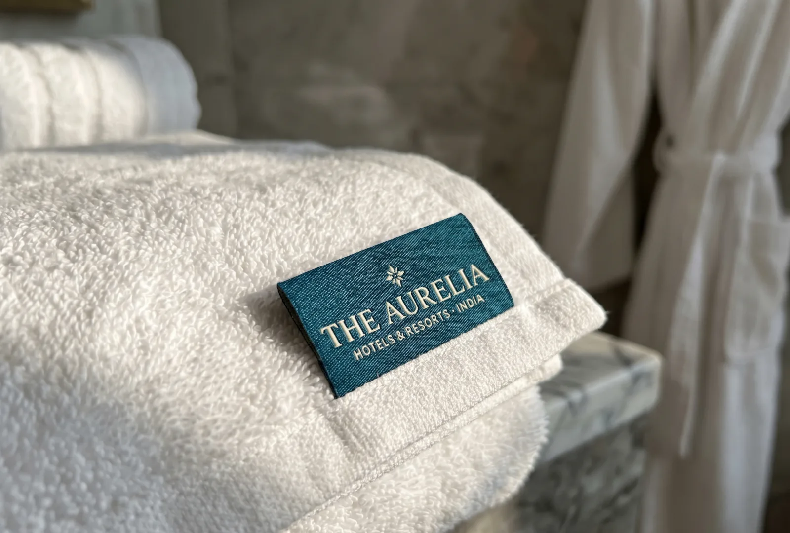 Deep teal woven label printed The Aurelia · Hotels & Resorts · India stitched onto a folded white hotel towel on a marble bathroom counter — example of an industrial-laundry-grade hotel linen label