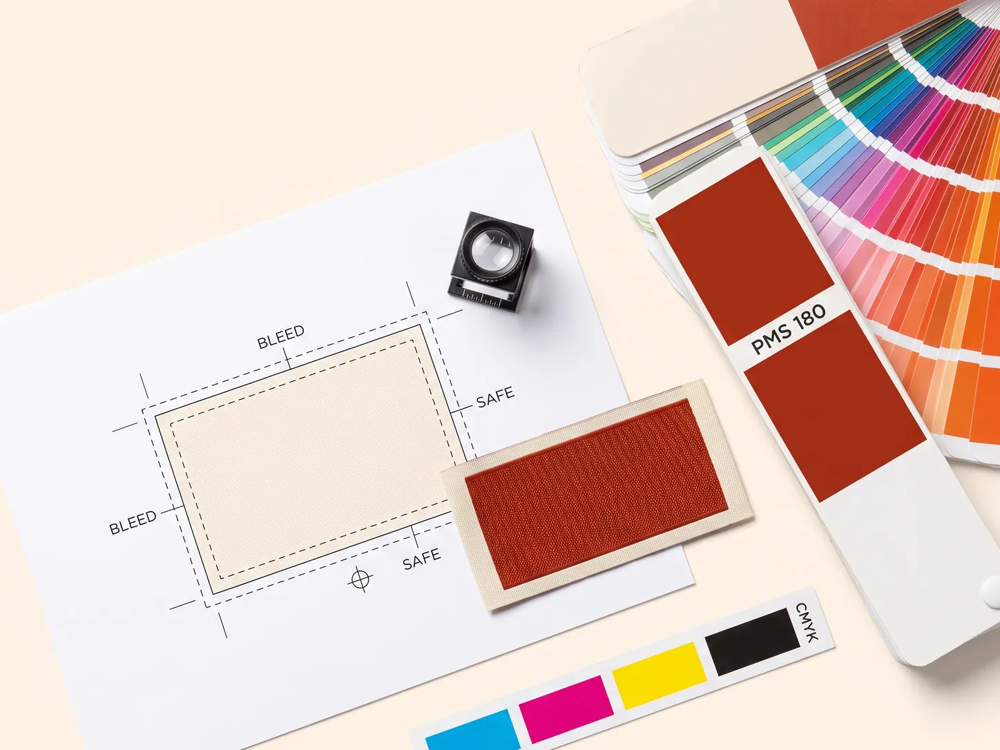 Label artwork print setup flat-lay: a proof sheet marked with bleed and safe lines and a registration target, a loupe, a woven label swatch, a PMS 180 Pantone fan deck and a CMYK colour strip