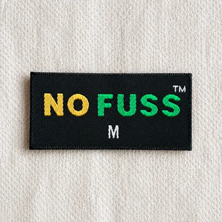 No Fuss black woven brand label with multi-colour yellow and green wordmark, TM mark and white M size marker, streetwear brand woven label sample