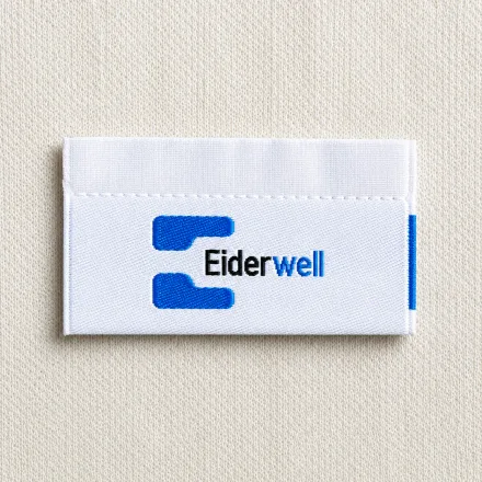 Eiderwell white centre-fold woven brand label with blue letter-E icon and italic wordmark, premium apparel woven neck label sample