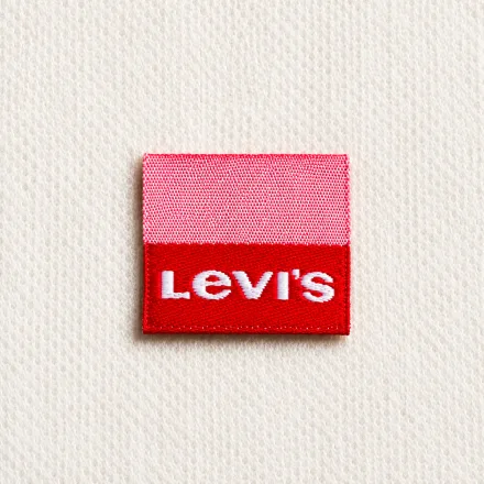 Small pink-and-red woven brand label sample with bold white block-letter wordmark: denim and casual-wear design reference, woven label finish