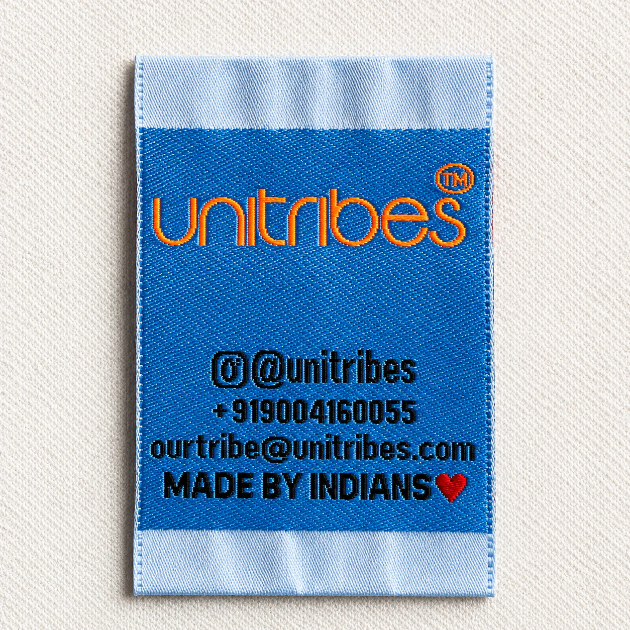 Unitribes blue satin printed brand label with orange wordmark, Instagram handle, phone, email and Made by Indians tagline — D2C brand satin printed label sample