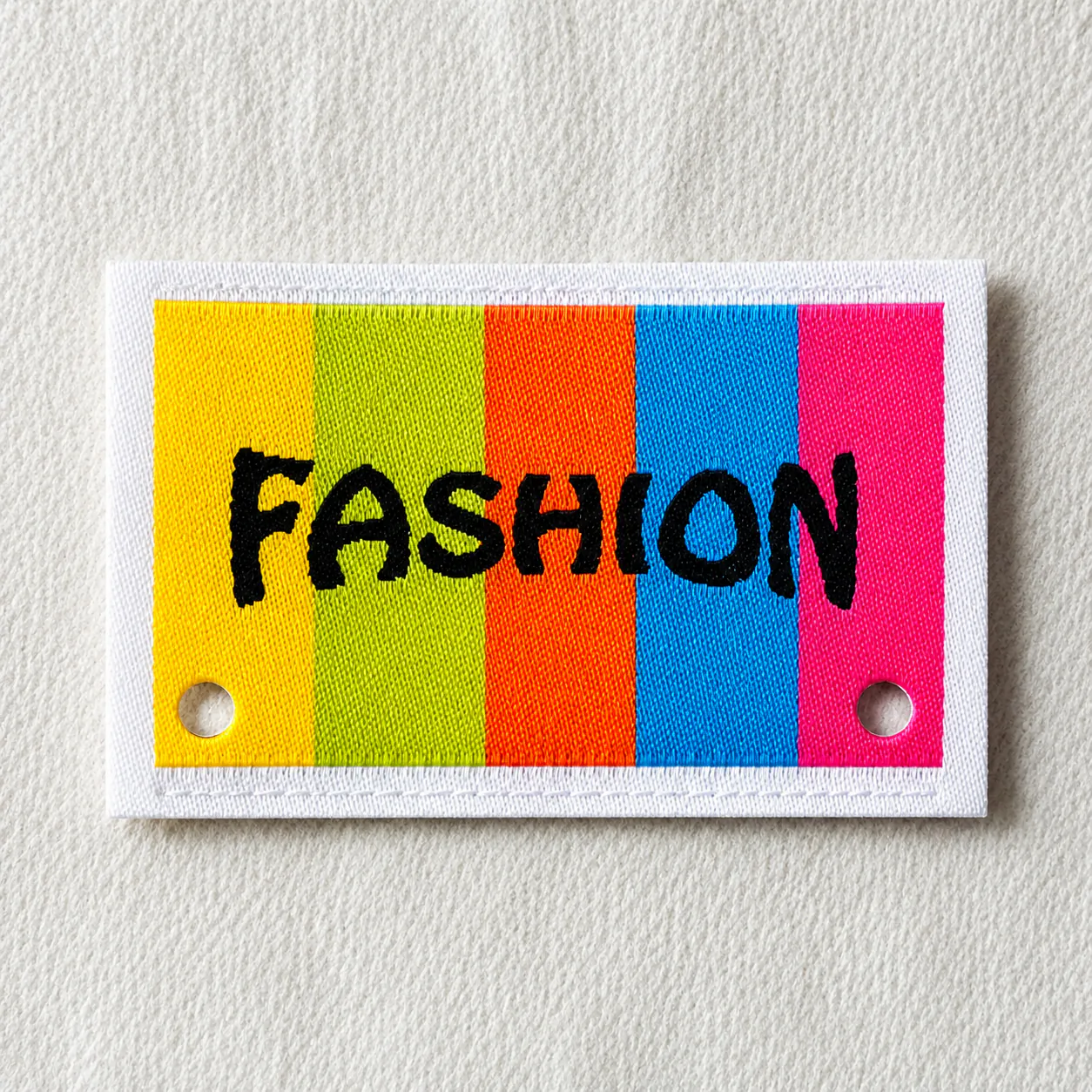 Multi-colour striped woven brand label with bold black hand-lettered FASHION wordmark and metal eyelets — vibrant streetwear woven brand label sample