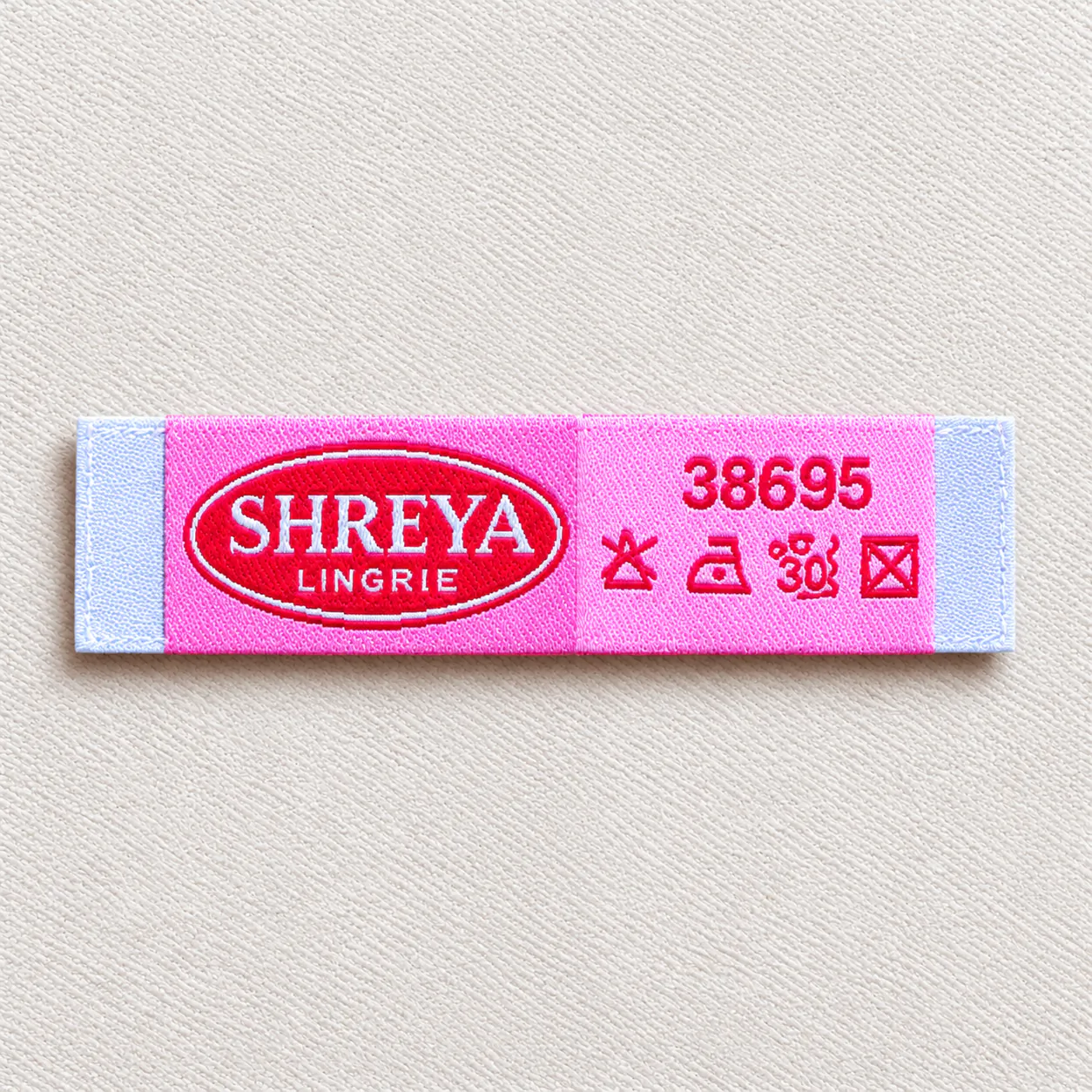 Shreya Lingrie pink-and-blue woven wash-care brand label with red oval logo, item code 38695 and red care-symbol panel — lingerie wash-care woven label sample