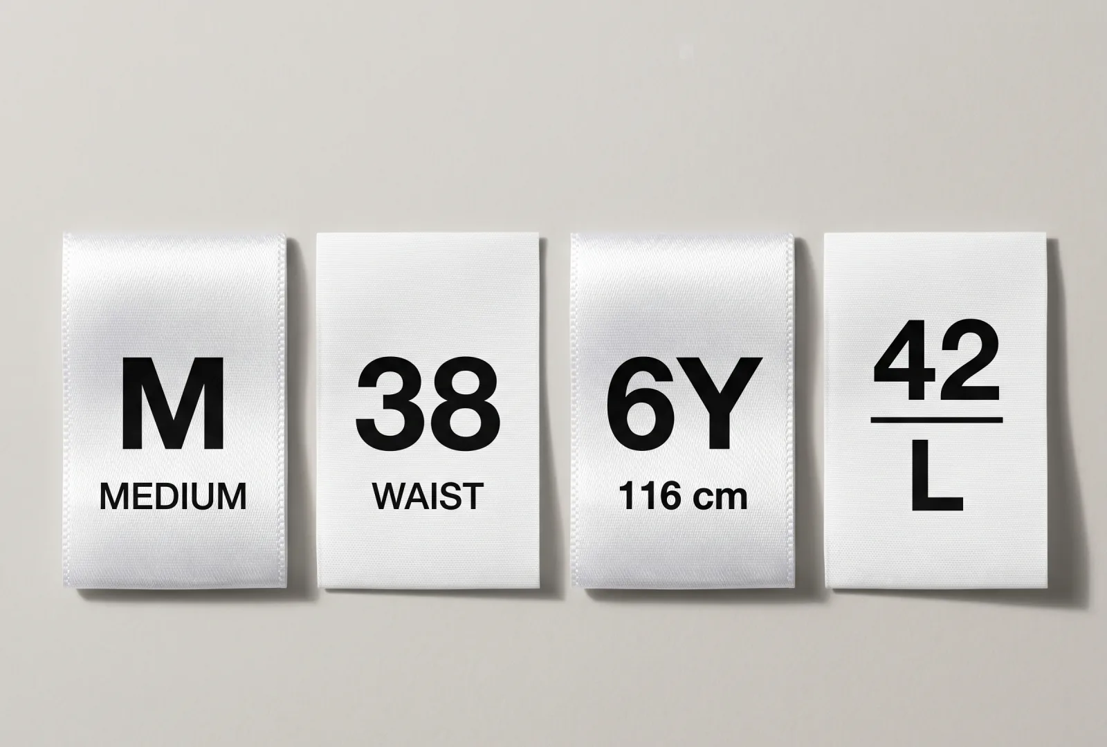 Four white satin size labels showing different sizing systems — alpha (M / Medium), waist measurement (38 / Waist), kids height (6Y / 116 cm) and EU-alpha dual (42 / L) — example of garment size labels for apparel brands