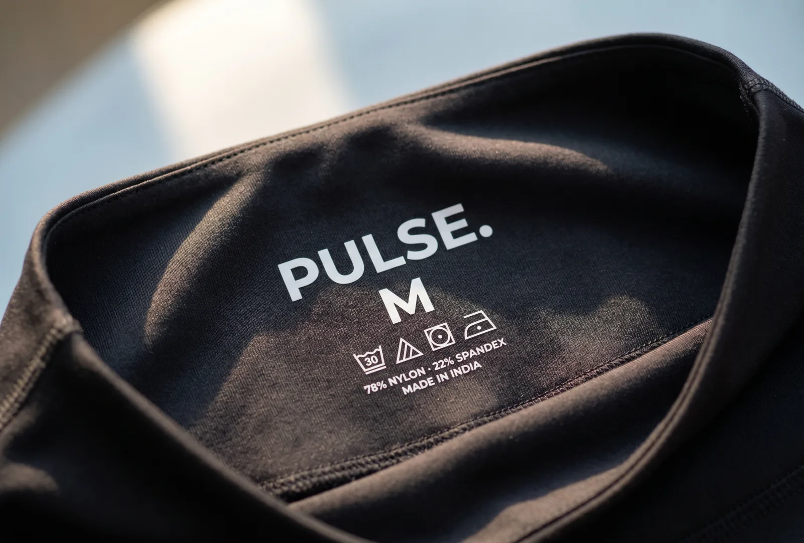 Inside neck of a black activewear top showing a white tagless heat-pressed print — PULSE. brand, size M, care symbols and 78% nylon 22% spandex Made in India — example of a tagless heat-press label for activewear